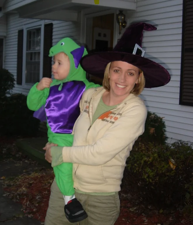 Becky Fontana in a purple witch hat, holding a toddler in a green dragon costume