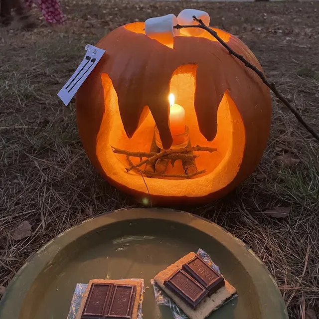 A campfire pumpkin with marshmallows on a stick, next to a plate of s’mores